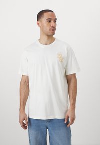 Vit bomulls t-shirt med korta ärmar, rund halsringning och ett gyllene tryck som visar "1956" på vänster bröst. Bärs med blå jeans.