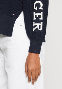 Marineblauer Strickpullover mit weißem "GER"-Text auf dem Ärmel, gerippten Bündchen und lockerer Passform. Kombiniert mit weißen Jeans.