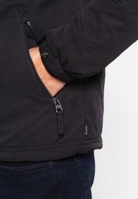 Czarna kurtka na suwak z ściągaczem przy rękawach i bocznymi kieszeniami. Posiada matowe wykończenie oraz małą metkę z logo w pobliżu dołu.