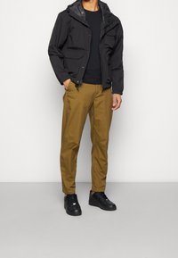 Černá lehká bunda s kapucí, kapsy na zip, v kombinaci s khaki kalhotami a černými teniskami. Hladká textura, moderní střih, funkční design.