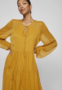 Vestido amarelo mostarda feito de tecido leve, com um decote de laço, mangas longas bufantes e um design de saia em camadas.