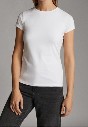Kvinde iført en simpel hvid slim fit t-shirt med korte ærmer og mørke, højttaljede jeans mod en neutral baggrund.