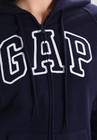 Sweat à capuche zippé bleu marine avec de grandes lettres blanches formant "GAP" sur le devant, en tissu texturé et avec des cordons de serrage sur la capuche.