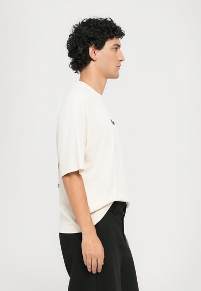 Camiseta de manga corta color crema con un ajuste relajado, cuello redondeado y detalle impreso en negro en la parte delantera. Combinada con pantalones negros.