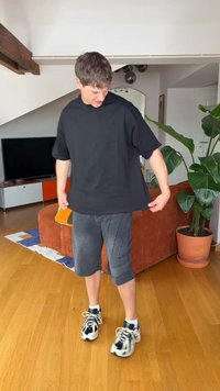 Άνδρας που ρυθμίζει υπερμεγέθη μαύρο T-shirt, στέκεται σε ξύλινο δάπεδο σε σαλόνι με πορτοκαλί καναπέ, μεγάλο πράσινο γλαστράκι και τηλεόραση στο φόντο.