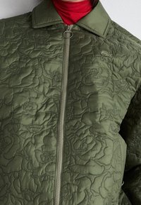 Kurtka w kolorze oliwkowym o pikowanym wzorze z zamkiem z przodu, z kwiatową teksturą, spiczastym kołnierzem i logo Lacoste na piersi. Czerwony golf pod spodem.