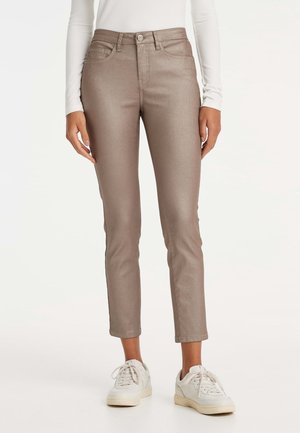 Schmal geschnittene metallisch braune Hose, getragen mit weißen Sneakern und einem weißen Langarmtop, gezeigt von der Taille bis zu den Füßen.