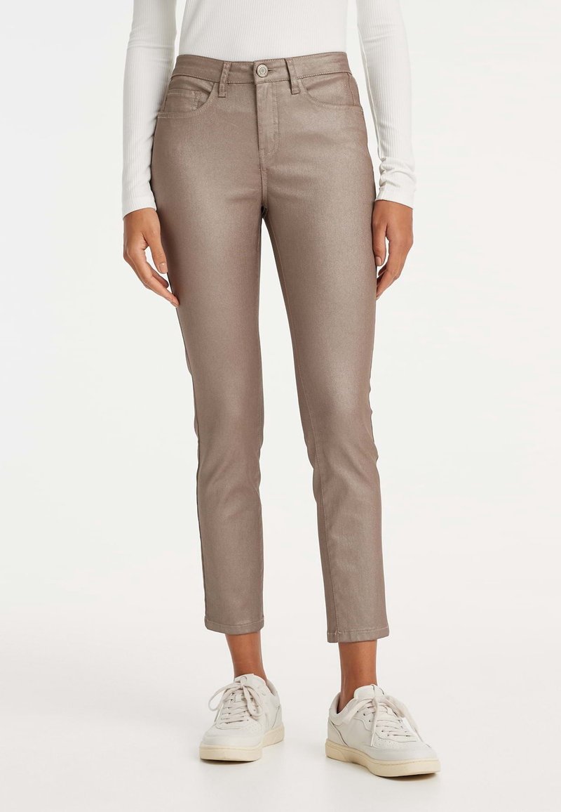 Pantalon slim marron métallisé porté avec des baskets blanches et un haut blanc à manches longues, visible de la taille jusqu'aux pieds.