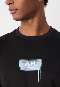 Camiseta de algodón negra con un gráfico frontal en azul claro que presenta el texto "AM" y "ANTONY MORATO" sobre un fondo de pinceladas texturizadas.