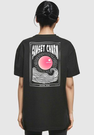 Mustmust valge trükk alaosas mustal T-särgil, millel on suur graafika seljal, kus on roosa päike, lained ja tekst "SUNSET CHARM" valges äärises. Lühikesed varrukad.