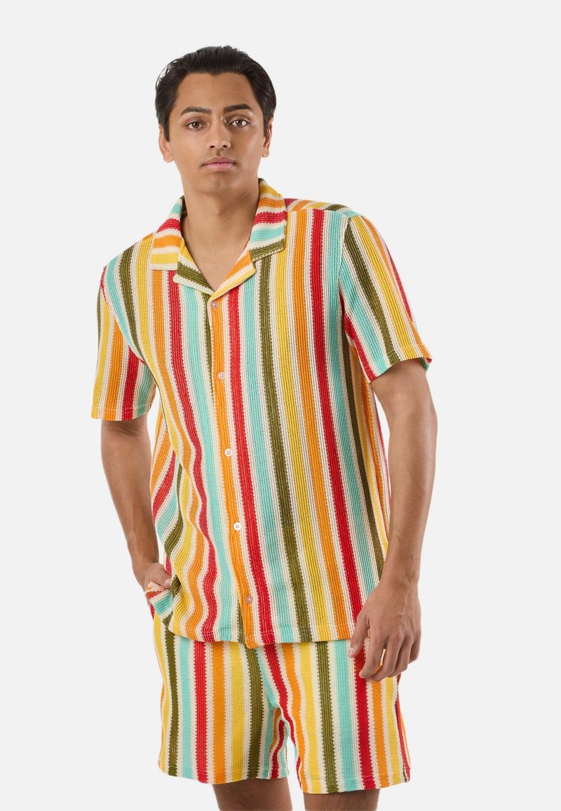 Homme portant une chemise à manches courtes à rayures verticales colorées et un short assorti, debout avec une main dans la poche.