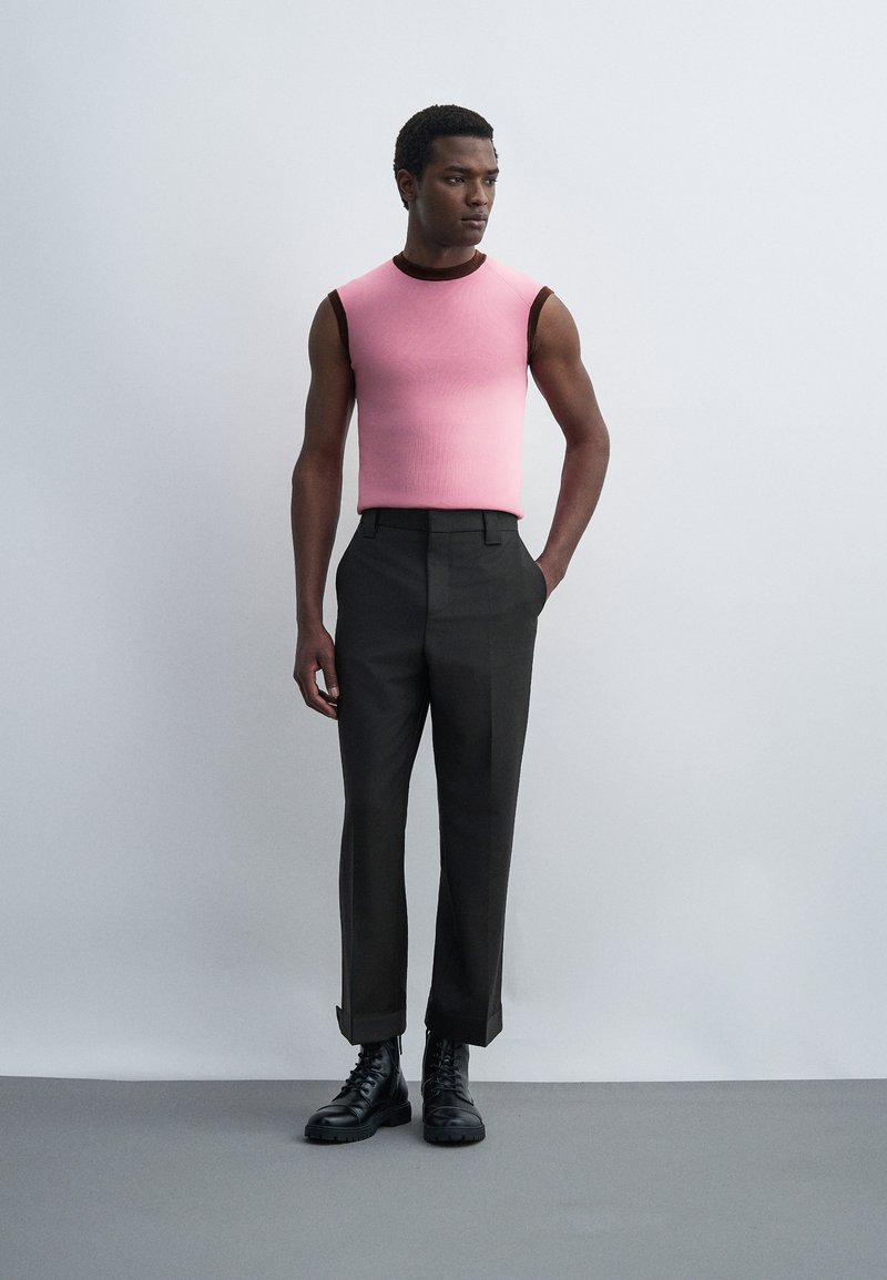 Man standing wearing sleeveless pink top with dark trim, black high-waist trousers, and black lace-up boots against plain wall.