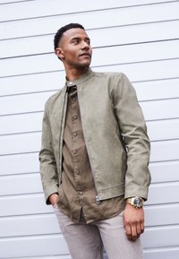 Veste en daim vert olive clair avec fermeture éclair et col montant, associée à une chemise marron à boutons et un pantalon beige clair.