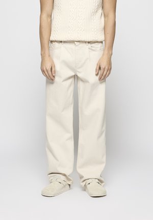 Personne portant un pantalon large écru, un pull en maille sans manches et des chaussures claires sans lacets, debout devant un fond uni.