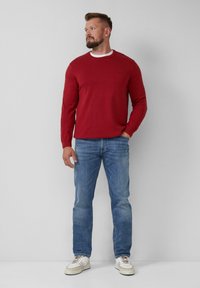 Roter Strickpullover mit Rundhalsausschnitt, kombiniert mit hellblauen Jeans und weißen Sneakern, vor einem grauen Hintergrund stehend.