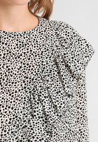 Blusa a pois bianca e nera con dettagli arricciati sulle spalle, caratterizzata da una vestibilità morbida e una texture di tessuto soffice.