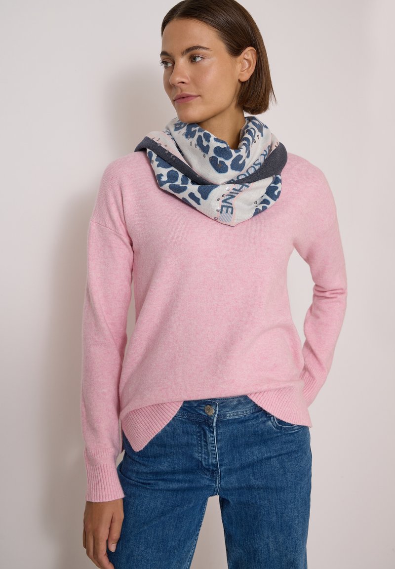 Femme portant un pull rose clair, un jean bleu et une écharpe blanche et bleue à motifs, debout, une main sur la hanche, regardant sur le côté.