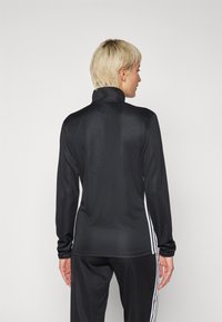 Svart langermet treningstopp med høy krage, laget av glatt stoff. Har hvite striper langs sidene og en tettsittende design.