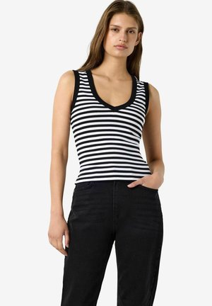 Jeune femme portant un haut sans manches rayé noir et blanc avec bordure noire et un pantalon noir taille haute, debout avec une main dans la poche.