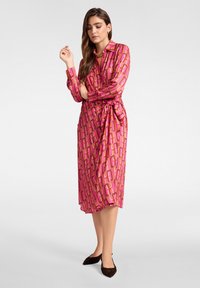 Vestido largo rosa de manga larga con un patrón geométrico, presenta un cuello con solapa y un lazo en la cintura, está hecho de una tela suave y se combina con zapatos planos oscuros.