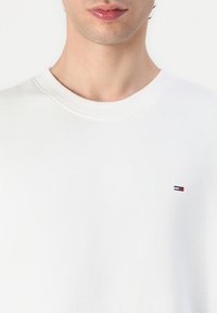 Hvit sweatshirt med rund hals og ribbede mansjetter. Har en liten brodert merkevarelogo i marineblått, rødt og hvitt på brystet. Myk tekstur.