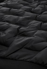 Černá prošívaná tkanina s hladkou texturou, která obsahuje řadu rovnoměrně rozmístěných výstupků vytvářejících vzor ve tvaru diamantů. Měkký a lehký vzhled.