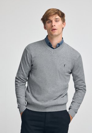 Suéter gris con un diseño de escote en V y puños acanalados, llevado sobre una camisa azul con cuello. Cuenta con un pequeño logotipo en el lado izquierdo del pecho.