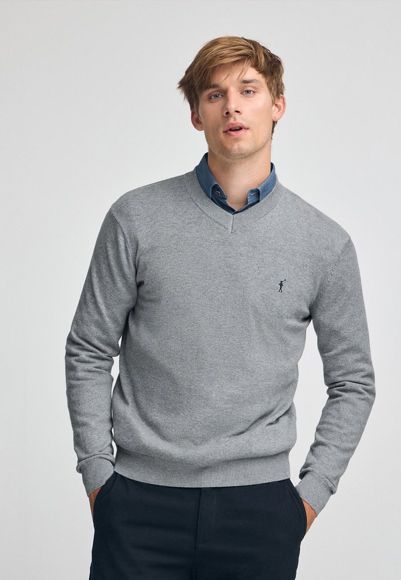 Maglione grigio con scollatura a V e polsini a coste, indossato sopra una camicia blu con colletto. Presenta un piccolo logo sul lato sinistro del petto.