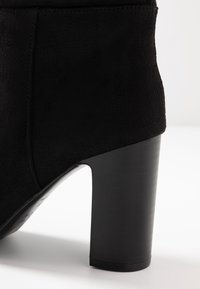 Bottine noire en daim avec un talon épais. Caractérisée par une texture lisse et des lignes épurées, mettant en avant un design moderne et minimaliste.