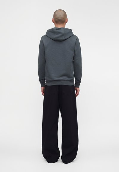 Personne portant un sweat à capuche gris et un pantalon noir ample, debout dos au spectateur sur un fond uni.