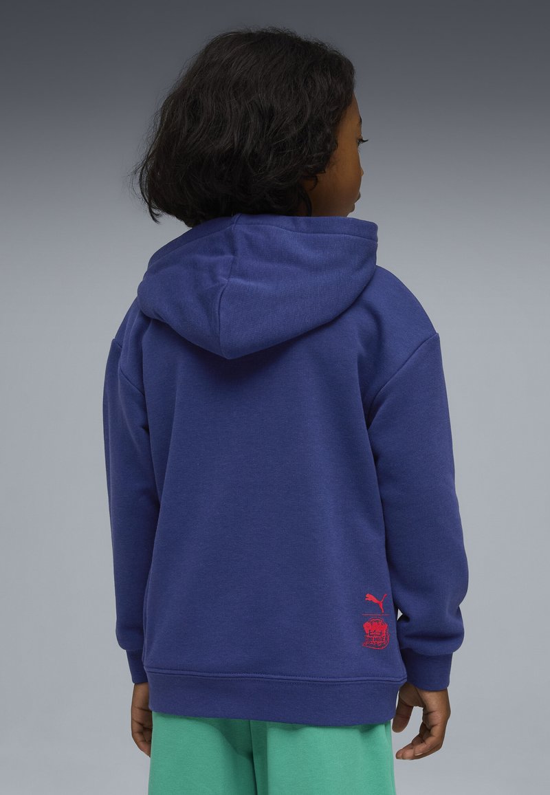 Enfant aux cheveux courts et bouclés portant un sweat à capuche bleu avec le logo rouge Puma, pantalon vert, de dos sur un fond gris.