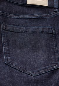 Un tissu en denim bleu foncé présente une poche arrière cousue avec un rabat courbé, une texture lisse et un léger brillant. Détail de l'étiquette visible au-dessus de la poche.