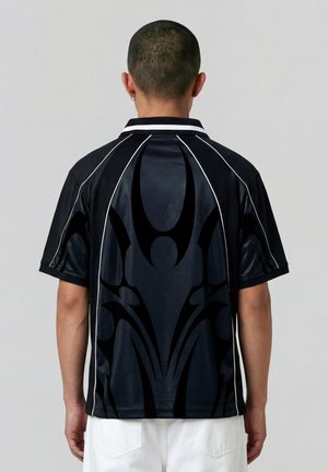 Mann med kort hår iført svart og hvit sportsskjorte med stort svart abstrakt motiv på ryggen og hvite bukser, stående mot en grå bakgrunn.