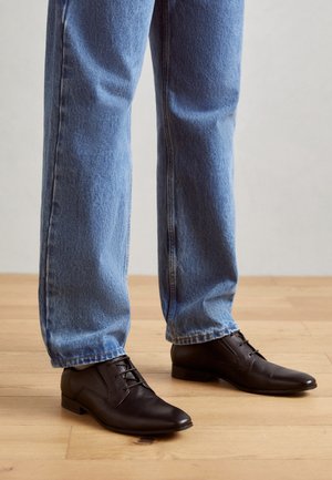 Scarpe di pelle nere con punta affusolata, design con lacci e tacco basso, indossate con jeans dritti blu chiaro su un pavimento di legno.