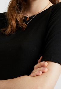 Černé tričko s krátkým rukávem z hladké tkaniny, nošené s vrstvenými zlatými náhrdelníky se sbíhajícími se obdélníkovými přívěsky, s překříženýma rukama.