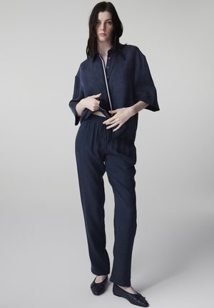 Kobieta stojąca, ubrana w luźne ciemnoniebieskie spodnie i pasującą zapinaną na guziki koszulę, poprawiająca koszulę, na jednolitym jasnoszarym tle.