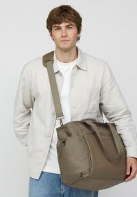 Jonge man met beige jas en wit overhemd, die een grote taupekleurige schoudertas met crossbody-riem vasthoudt tegen een effen achtergrond.
