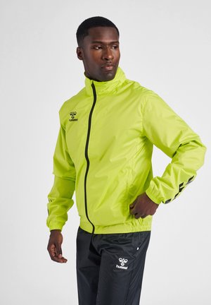 Heldere neon gele windbreaker jas met een ritssluiting aan de voorkant, elastische manchetten en zwarte accenten op de mouwen en het helmlogo. Gepaard met zwarte broek.