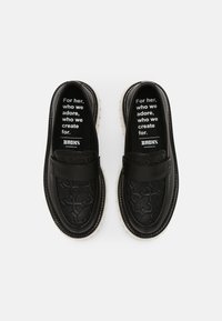 Une paire de mocassins en cuir noir avec un motif texturé et des semelles blanches, le texte à l'intérieur indique "Pour elle, que nous adorons, pour qui nous créons," marque Bronx.
