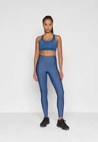 Αθλητικό σετ που περιλαμβάνει ένα εφαρμοστό σκούρο μπλε αθλητικό σουτιέν και ψηλόμεσες κολάν που ταιριάζουν, και τα δύο φτιαγμένα από ελαστικό ύφασμα. Μαύρα αθλητικά παπούτσια.