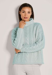 Frau, die einen hellblauen, grobstrickigen Rollkragenpullover mit langen Ärmeln trägt und vor einem einfarbigen, hellen Hintergrund steht.