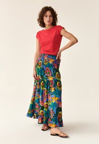 Haut rouge à manches courtes associé à une longue jupe fleurie colorée avec des motifs bleus, verts, jaunes et orange. Sandales à fines brides.