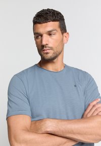 Lichtblauwe t-shirt met korte mouwen en een ronde halslijn, voorzien van een subtiele textuur. Kleine zwarte accentdetails op de borst. Gladde stof.