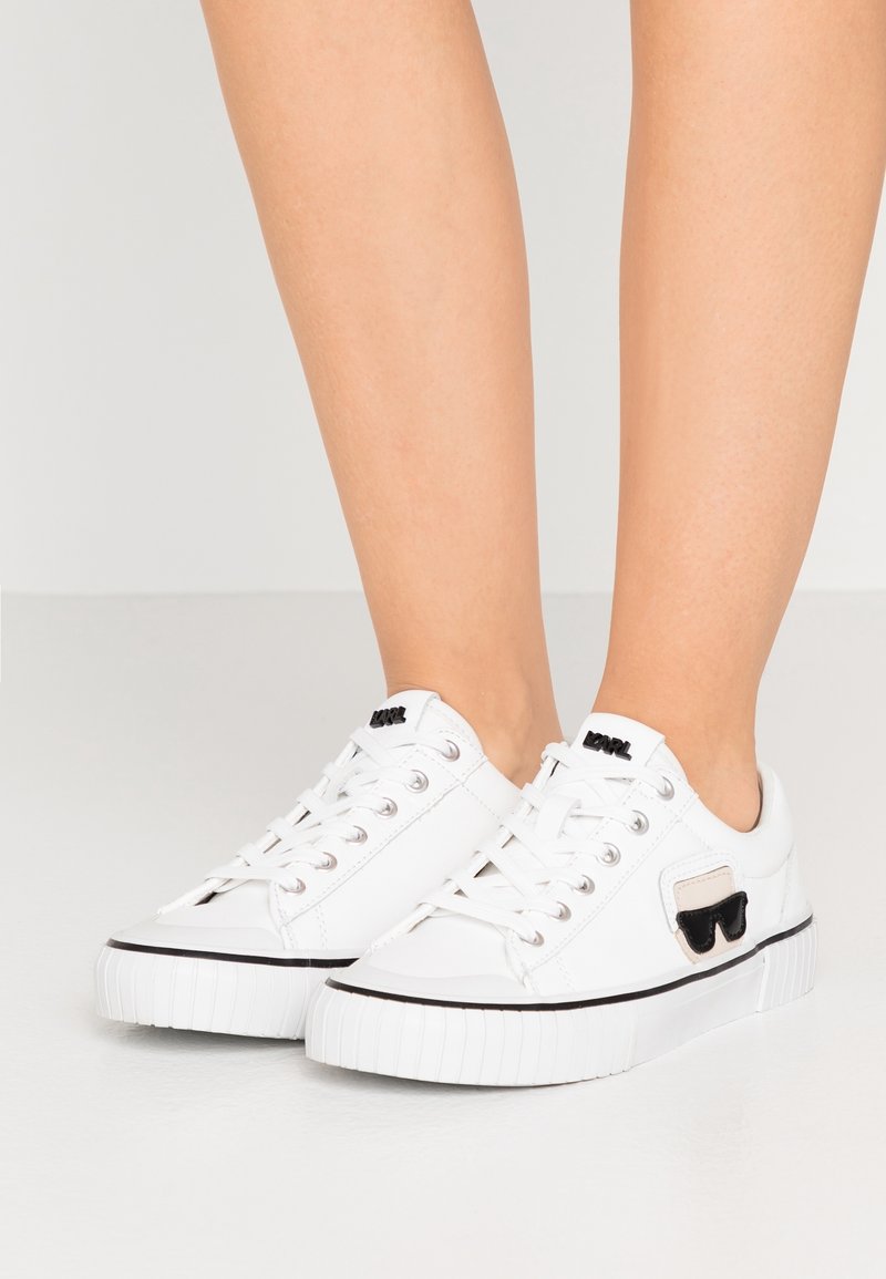 Weiße Low-Top-Sneaker mit schwarzem Sonnenbrillen-Logo an der Seite, weißen Schnürsenkeln und schwarzem Rand, getragen von einer Person, die auf einem hellen Boden steht.