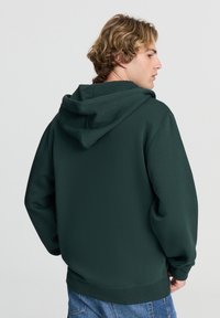 Felpa con cappuccio verde scuro, dalla texture morbida, vestibilità ampia e polsini a coste. Presenta una tasca a marsupio e un cappuccio regolabile con cordino.
