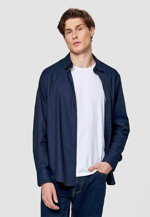 Młody mężczyzna z jasnobrązowymi włosami, ubrany w białą koszulkę pod rozpiętą granatową koszulę i ciemnoniebieskie dżinsy, stoi z ręką w kieszeni.