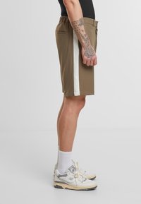 Shorts marron avec une bande blanche sur le côté, taille élastique et deux poches arrière. Portés avec des chaussettes blanches et des baskets. Tatouage détaillé visible.