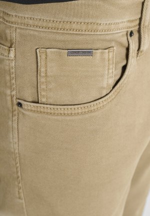 Close-up van tan denim broeken met gedetailleerde stiksels en een klein voorvak met een metalen label.