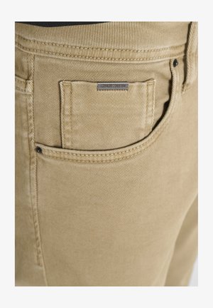 Close-up van tan denim broeken met gedetailleerde stiksels en een klein voorvak met een metalen label.