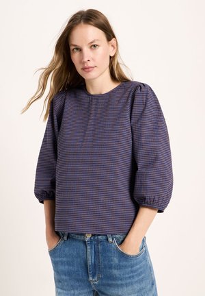 Woman with straight light brown hair wearing a blue and black checkered blouse with puffed sleeves and blue jeans with hands in pockets.
